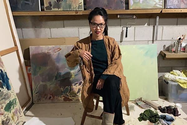 Andrea Bolima, wearing glasses and a brown coat, sits on a chair in an art studio surrounded by abstract paintings and art supplies.