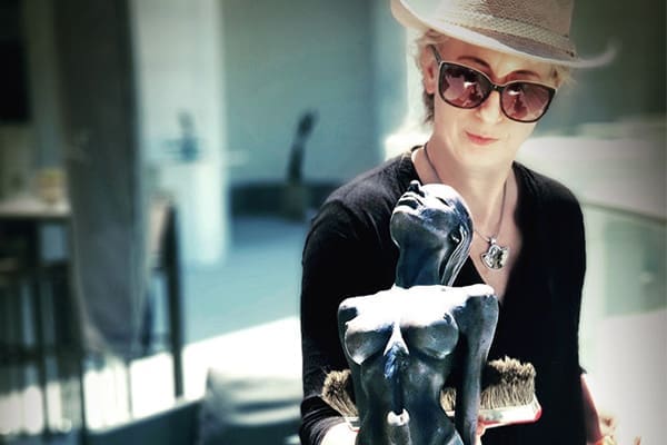 Anneke Bester, wearing a hat and sunglasses, behind a dark-colored, head-tilted female torso sculpture outdoors.