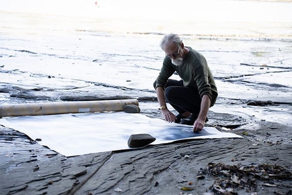 Garry Currin | The Art Precinct Garry Currin kneels on wet sand by the shoreline, arranging a large white sheet of paper held down with rocks.