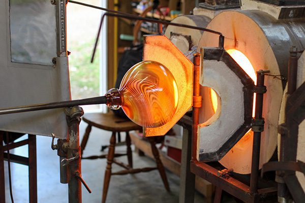 Justin Culina (out of frame) shapes molten glass with a blowpipe near a furnace in his glassblowing studio.