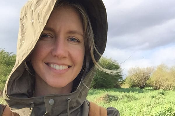Kate van der Drift stands outdoors in a grassy area, wearing an olive hooded jacket with cloudy skies and trees in the background.