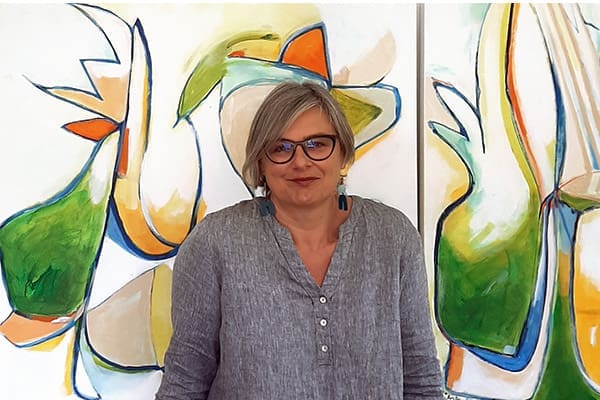 Kirsty Black, an artist with short gray hair and glasses, stands smiling in front of a colorful abstract painting, wearing a gray shirt.