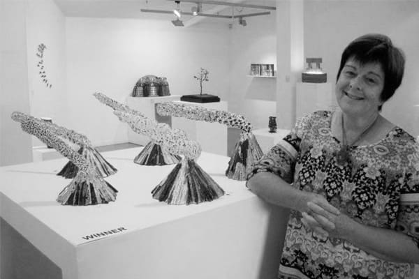 Artist Liz Sharek stands beside a winning abstract sculpture displayed on a pedestal in an art gallery.