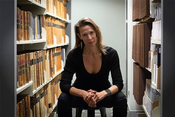 Mickey Smith, an artist with shoulder-length hair, sits on a stool between shelves filled with books and files in a library or archive.