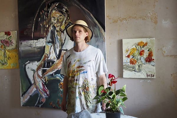 Nick Herd, dressed in a paint-stained shirt and straw hat, stands before abstract paintings with a potted plant on the table nearby.