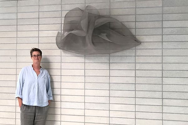 Artist Sally Smith stands beside her wall-mounted abstract wire sculpture on a light-colored brick wall.