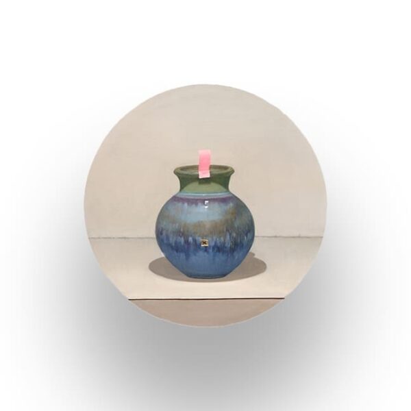 A round blue and green ceramic vase sits on a beige surface with a small pink rectangular object placed in its opening.