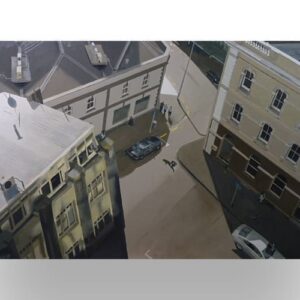 Aerial view of a quiet urban intersection with three buildings, a few people walking, and several parked cars on the street below.