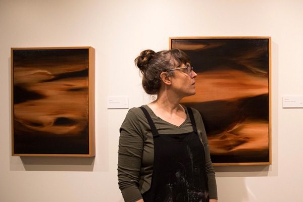 Hana Carpenter, wearing glasses, stands in front of two abstract paintings featuring dark brown and orange tones, gazing to her left.