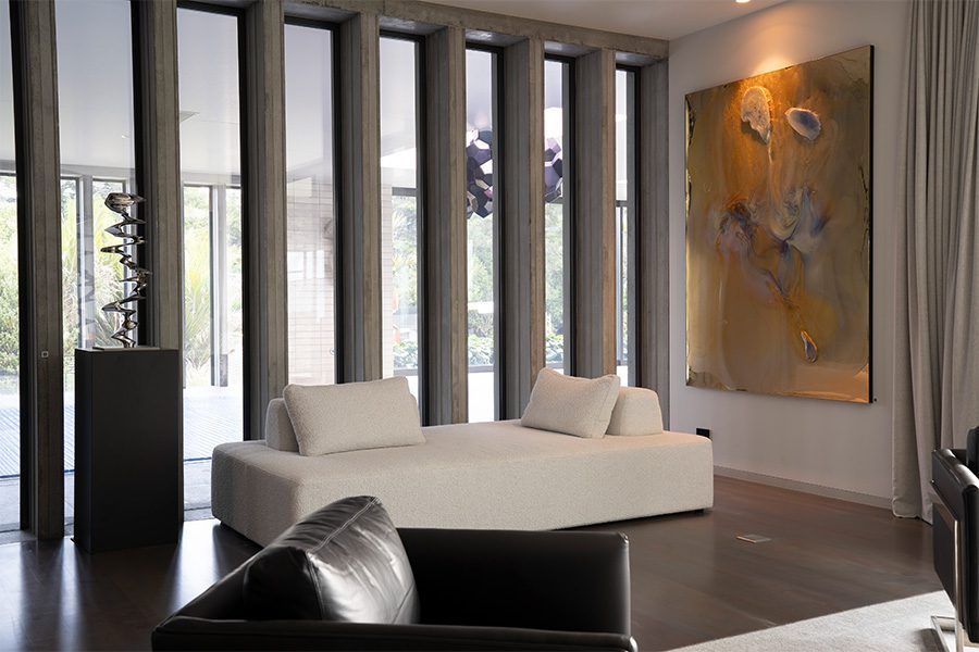 A modern living space with a comfy white day bed and a leather armchair, wall art of an abstract painting, a silver sculpture sits upon a podium, through the windows a large geometric sculpture hangs from the ceiling.