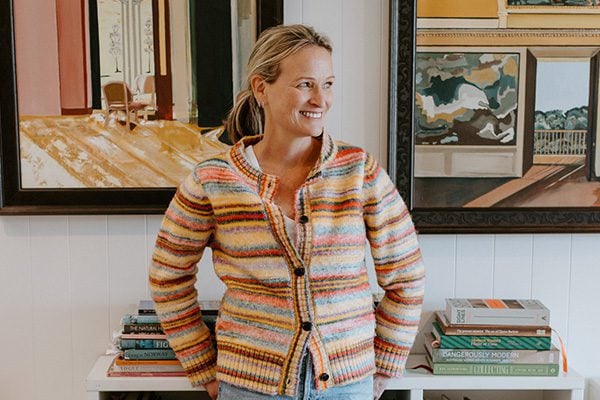 Tori Beeche, artist, leans against a bookcase, two of her framed paintings hang on the wall behind her, she's wearing a cosy colourful knitted cardigan and jeans, her hair is in a ponytail, she smiles off to the right of the image.